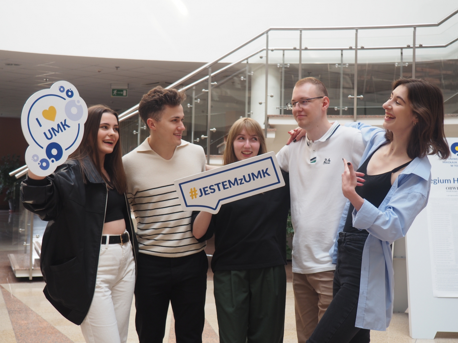 Grupa studentów pozuje w holu uczelni, trzymając tablice z napisami 'I love UMK' i 'Jestem z UMK', tworząc swobodną, radosną scenę promującą kierunek architektura informacji.