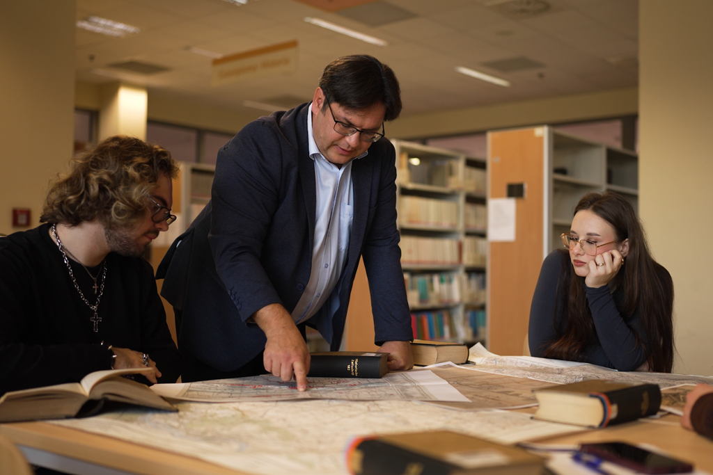 Podczas zajęć z historii osoba prowadząca przy stole w bibliotece wskazuje na mapę, a dwie osoby siedzące obok analizują materiały źródłowe.
