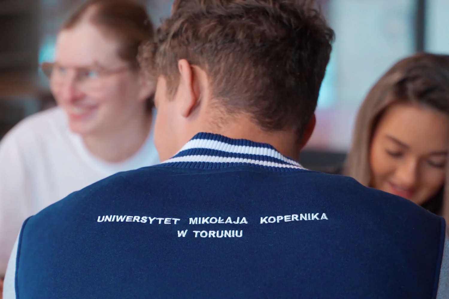 Student siedzi w bluzie z napisem Uniwersytet Mikołaja Kopernika w Toruniu na tle uśmiechniętych studentek.