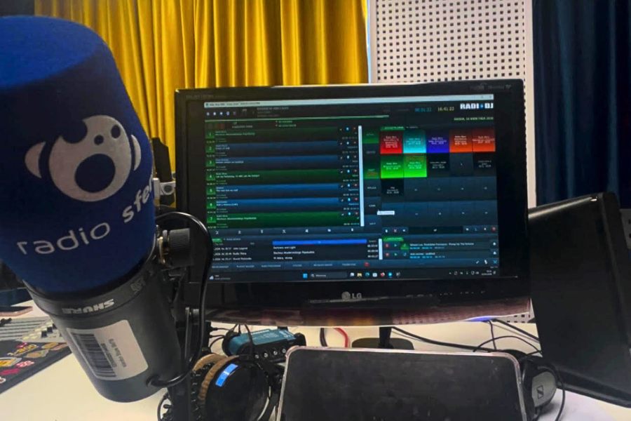 Na zdjęciu widać stanowisko radiowe z mikrofonem ?Radio Sfera?, słuchawkami oraz monitorem z oprogramowaniem do emisji audycji. W tle znajdują się zasłony i panele akustyczne obecne w studiu nagraniowym.