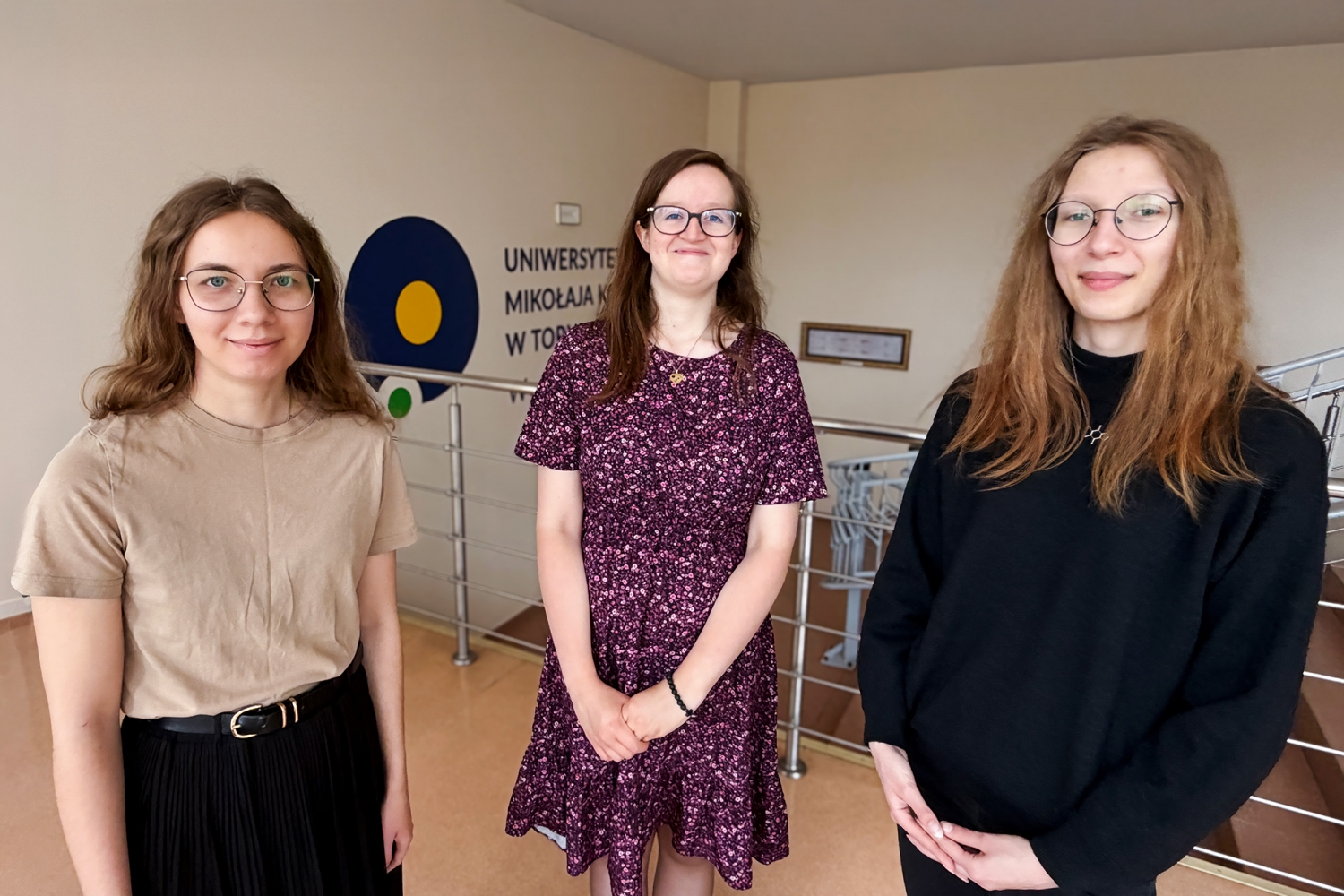 Na zdjęciu są studentki nauk o rodzinie na korytarzu Wydziału Teologicznego UMK przy ul. Gagarina 37 w Toruniu. Trzy studentki stoją blisko aparatu. Po lewej stronie znajduje się studentka w beżowej koszulce i okularach. W środku stoi studentka w bordowej sukience we wzory, z dłońmi złożonymi przed sobą. Po prawej stronie stoi studentka w czarnym swetrze i okularach. W tle widoczna jest metalowa balustrada, fragment logo Wydziału Teologicznego UMK oraz napis na ścianie.