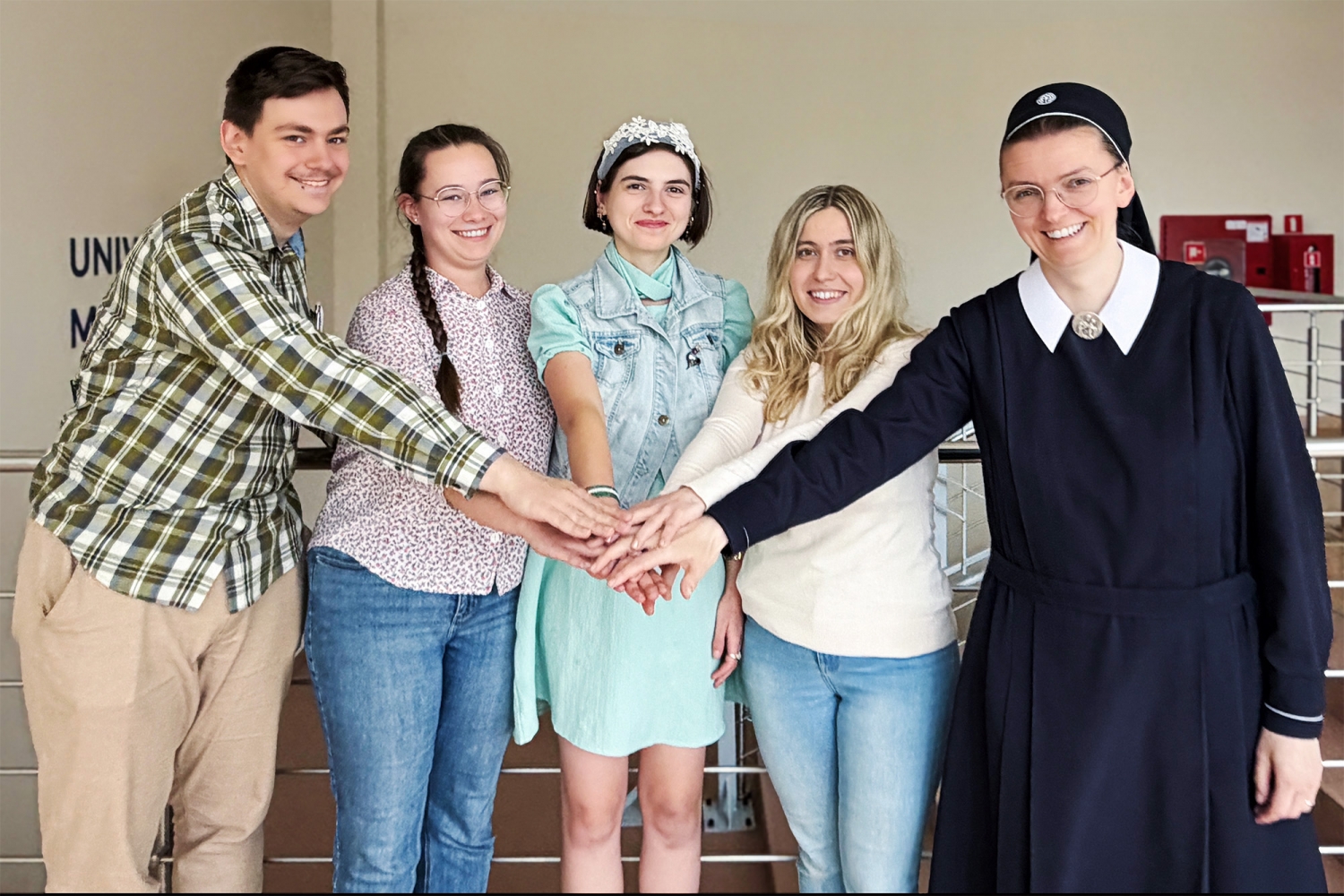 Na zdjęciu są studenci teologii na korytarzu Wydziału Teologicznego UMK przy ul. Gagarina 37 w Toruniu. Pięcioro studentów stoi blisko siebie i układa dłonie razem na środku kadru. Po lewej stronie stoi młody mężczyzna w koszuli w kratę. Obok niego stoi młoda kobieta w jasnej koszuli i okularach. W centrum znajduje się kobieta w miętowej sukience i jasnej kamizelce. Po jej prawej stronie stoi blondynka w białym swetrze. Po prawej stronie kadru stoi siostra zakonna w granatowym habicie. W tle widać balustradę oraz wnętrze Wydziału Teologicznego UMK.