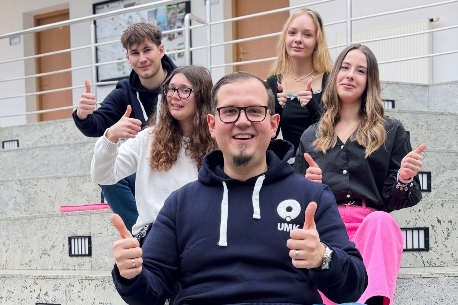 Pięć osób siedzi na schodach wewnątrz budynku uniwersyteckiego i pokazuje gest uniesionego kciuka.