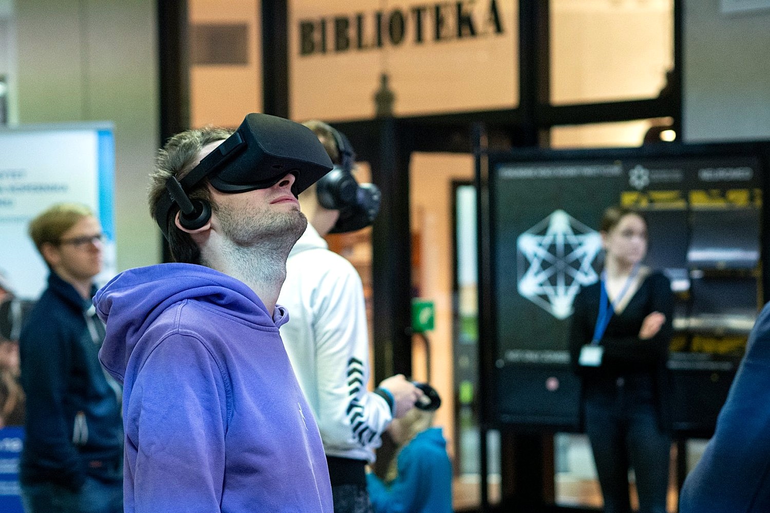 Mężczyzna w fioletowej bluzie z kapturem testuje gogle VR (wirtualnej rzeczywistości), patrząc w górę na tle wejścia do biblioteki, podczas gdy w tle inne osoby obserwują sytuację lub również korzystają ze sprzętu.