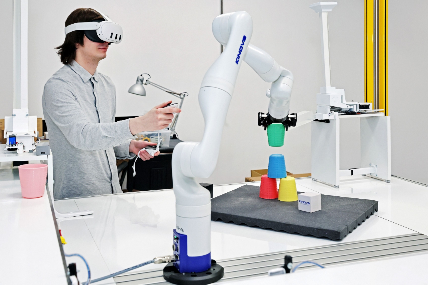 Tekst alternatywny: Pracownik w goglach rzeczywistości wirtualnej (VR, Meta Quest 3), który, trzymając kontrolery ruchowe, steruje w czasie rzeczywistym ramieniem robotycznym Kinova Gen3. Robot chwyta zielony kubek i ustawia go na piramidzie złożonej z trzech kolorowych kubków. W tle widoczne są podajniki z zasobnikami oraz taśmociąg. Scena ilustruje połączenie technologii VR z robotyką przemysłową ? jednego z dynamicznie rozwijających się obszarów badawczych związanych ze sztuczną inteligencją (AI).