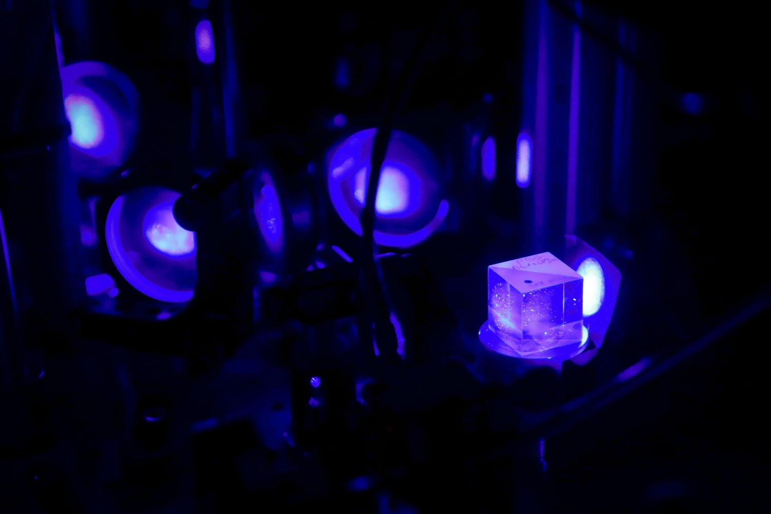 Wnętrze laboratorium Fizyki Atomowej, Molekularnej i Optycznej UMK skąpane w niebiesko-fioletowym świetle laserowym. Na pierwszym planie przezroczysta szklana kostka (element optyczny) oświetlona wiązką lasera. W tle szereg okrągłych soczewek lub luster w metalowych oprawach, przez które przechodzi ta sama wiązka światła.