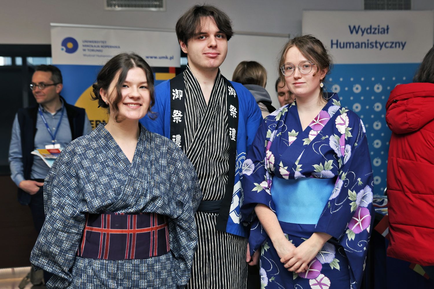 Dwie studentki i student japonistyki ubrani w kimona pozują przed banerem Wydziału Humanistycznego UMK.