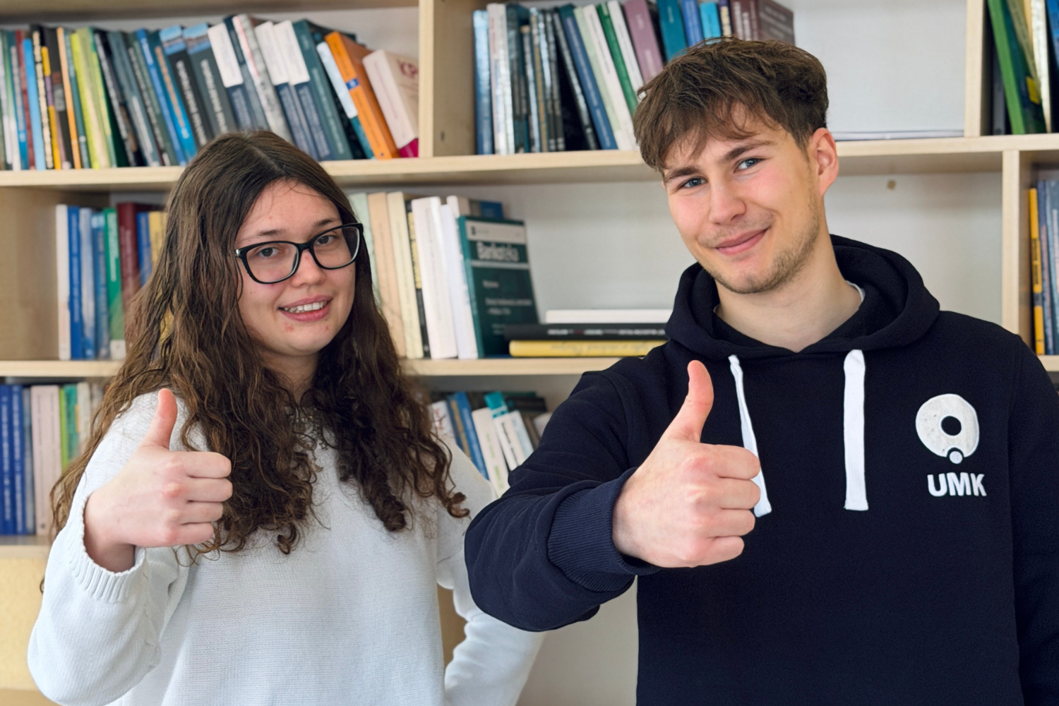 Studentka i student stoją przed regałem z książkami i pokazują gest uniesionego kciuka.