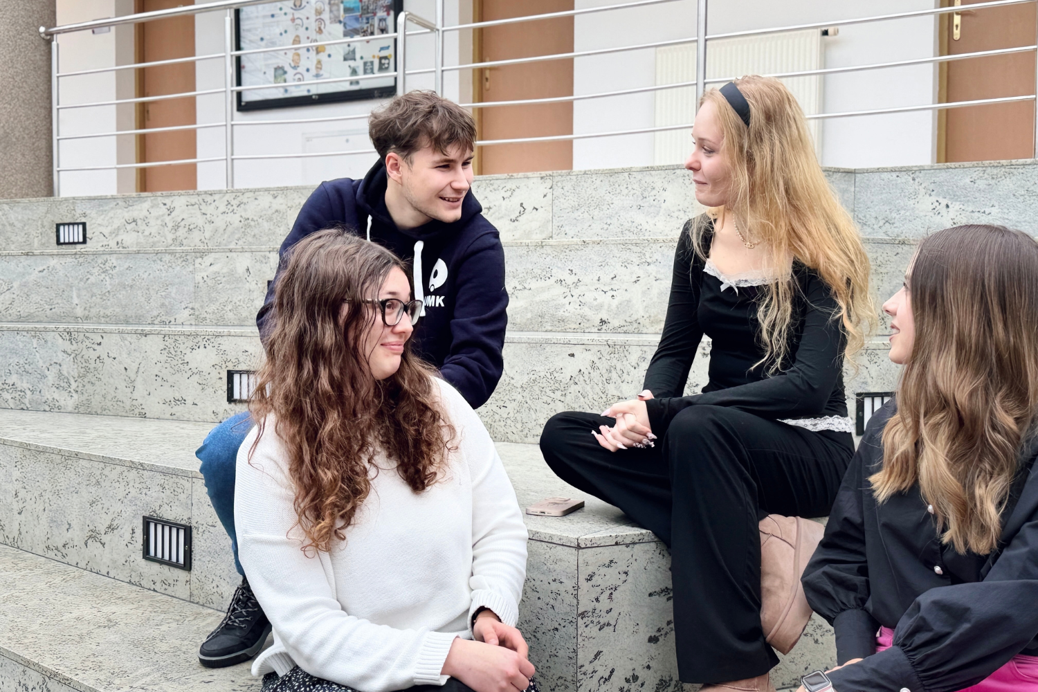 Czworo studentów siedzi na schodach uczelni i prowadzi rozmowę.