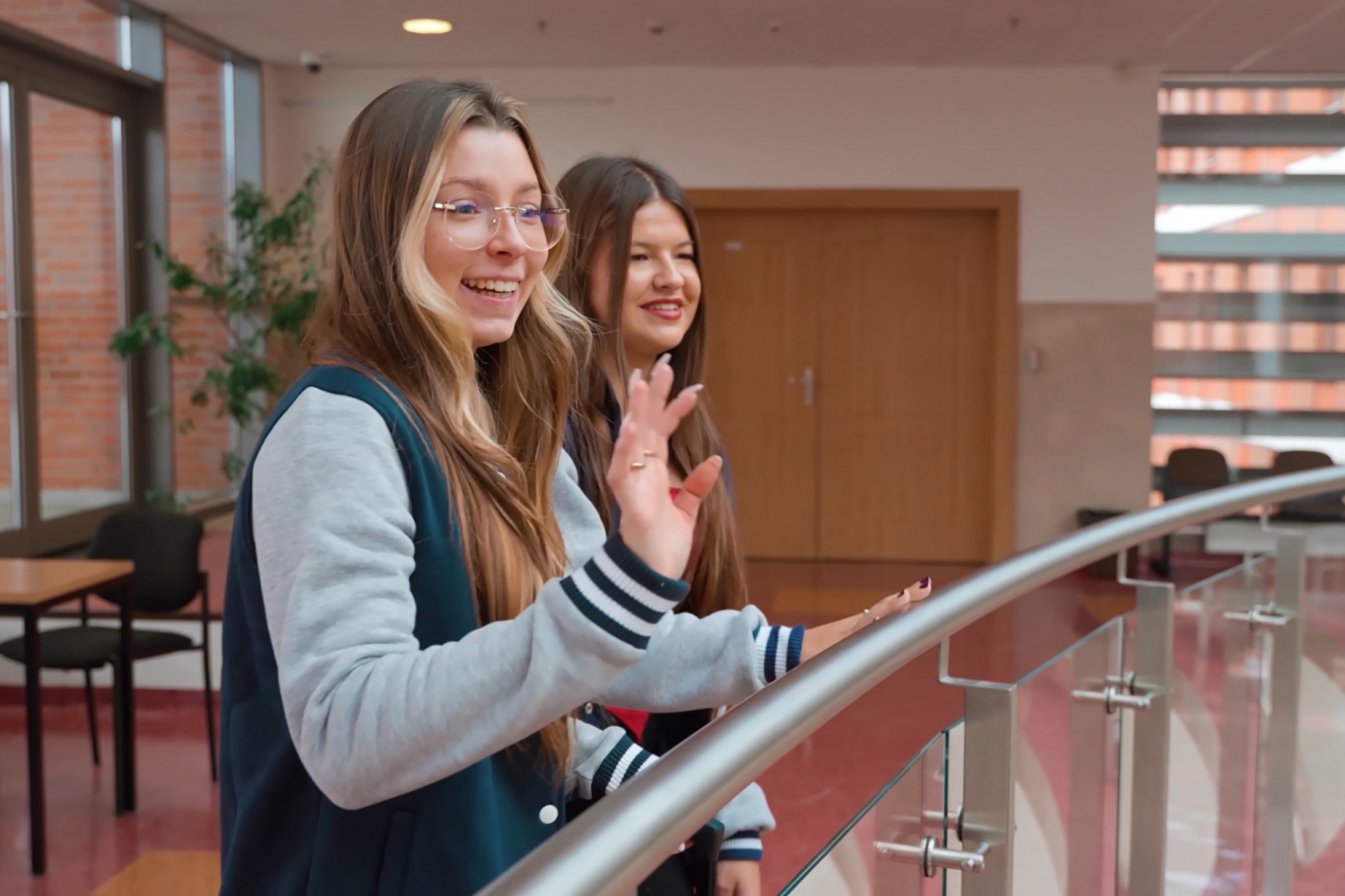 Dwie uśmiechnięte studentki pozdrawiają z zza balustrady.