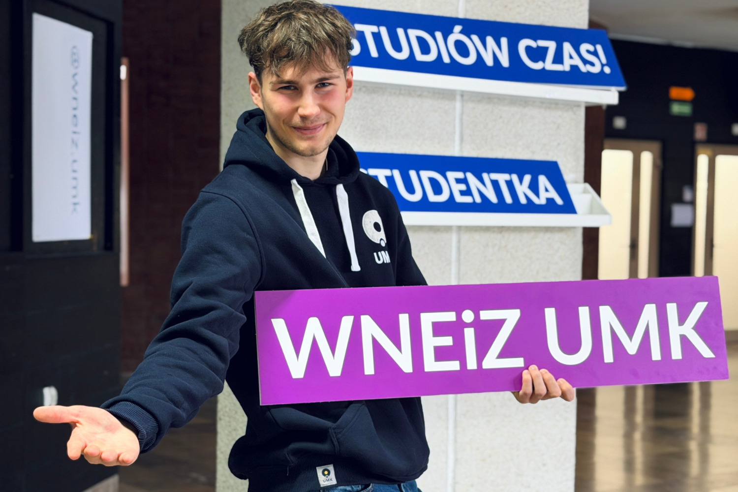 Student trzyma tablicę z napisem 'WNEiZ UMK' i gestem dłoni zachęca do zapoznania się z ofertą studiów.