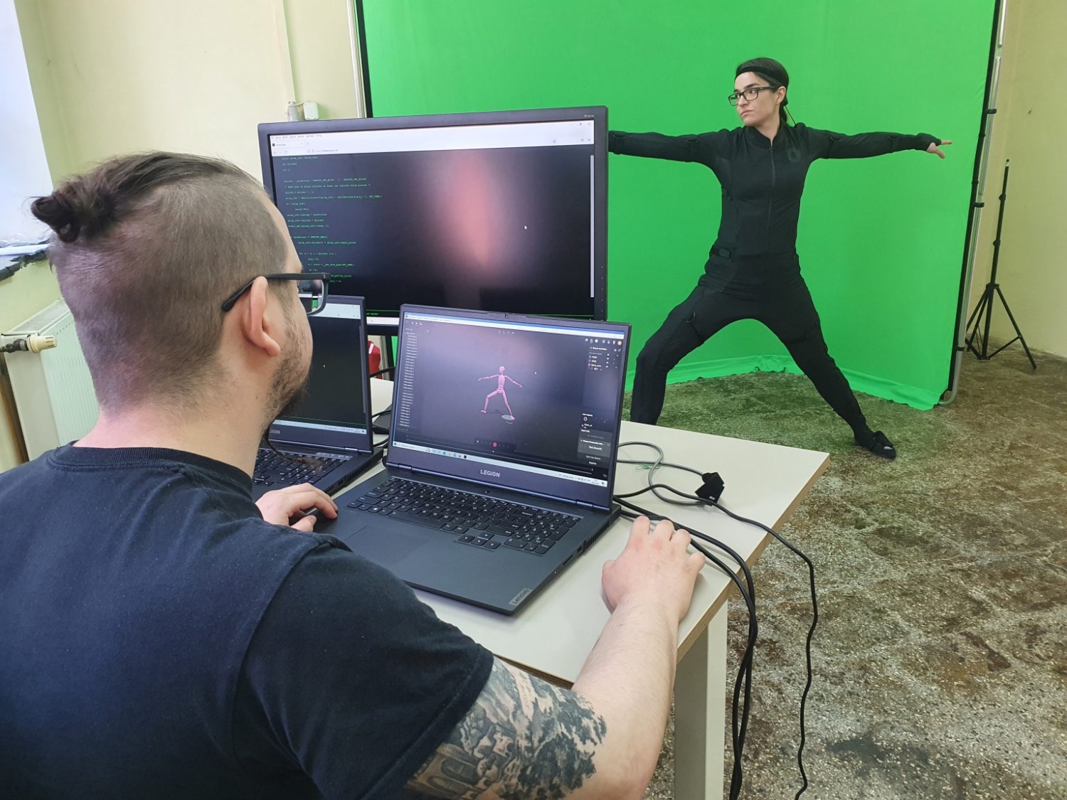 Mężczyzna pracuje przy komputerach nad projektem motion capture, podczas gdy kobieta pozuje na tle green screena. Na monitorach widoczny jest model postaci i kod programu do animacji ruchu.