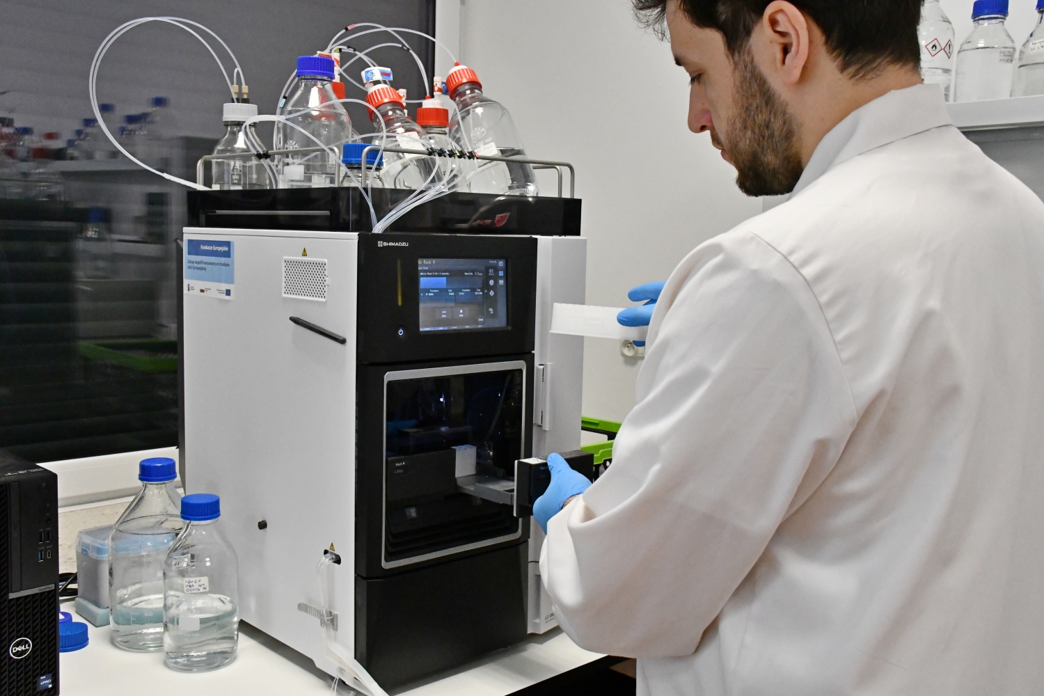 Badacz w białym fartuchu i niebieskich rękawiczkach obsługuje nowoczesny chromatograf (analizator laboratoryjny)  z podłączonymi butelkami z odczynnikami.