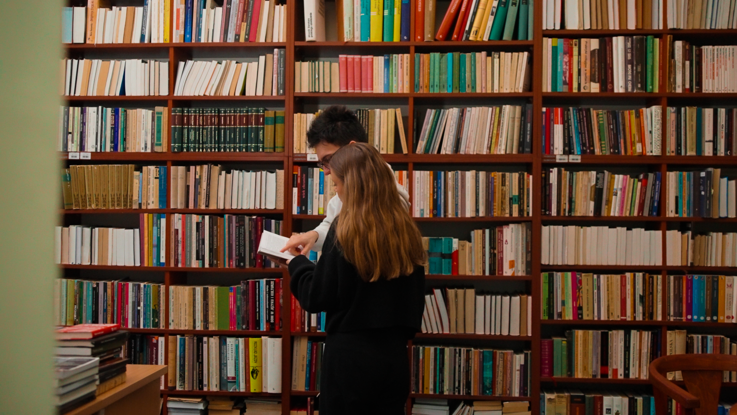 Dwoje studentów przegląda książkę w bibliotece przed regałami wypełnionymi publikacjami naukowymi.